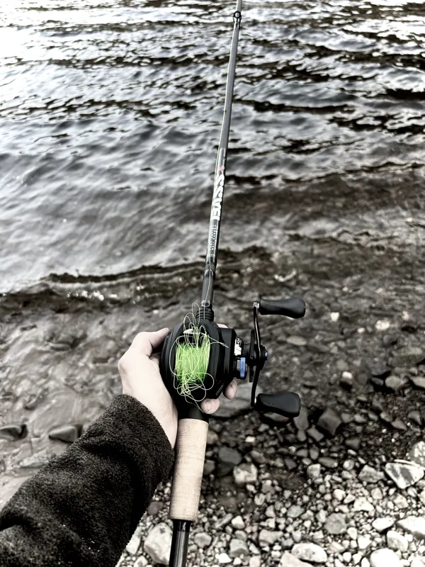 A hand holds a baitcasting reel over rocky water, loose green line looped around the spool