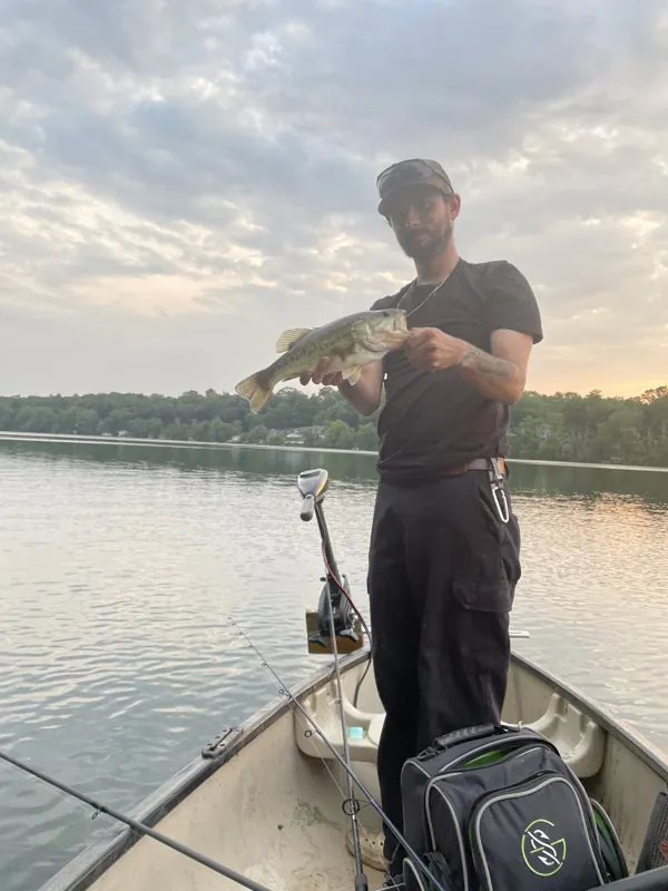 An angler stands in a boat on calm water at dusk, holding up a bass