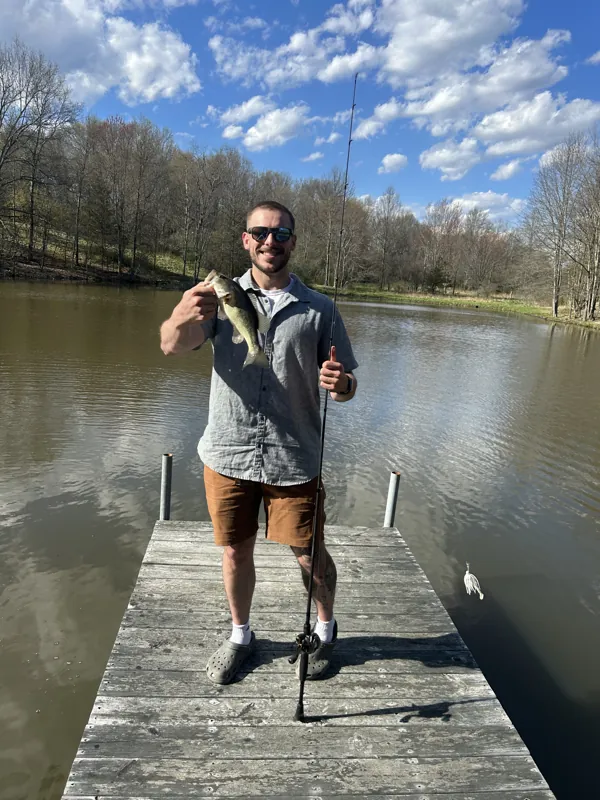 An angler on a wooden dock holds up a bass in one hand and a rod in the other