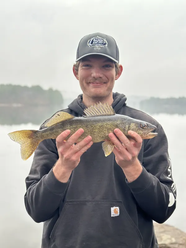 An angler holds a walleye in both hands in front of a misty lake
