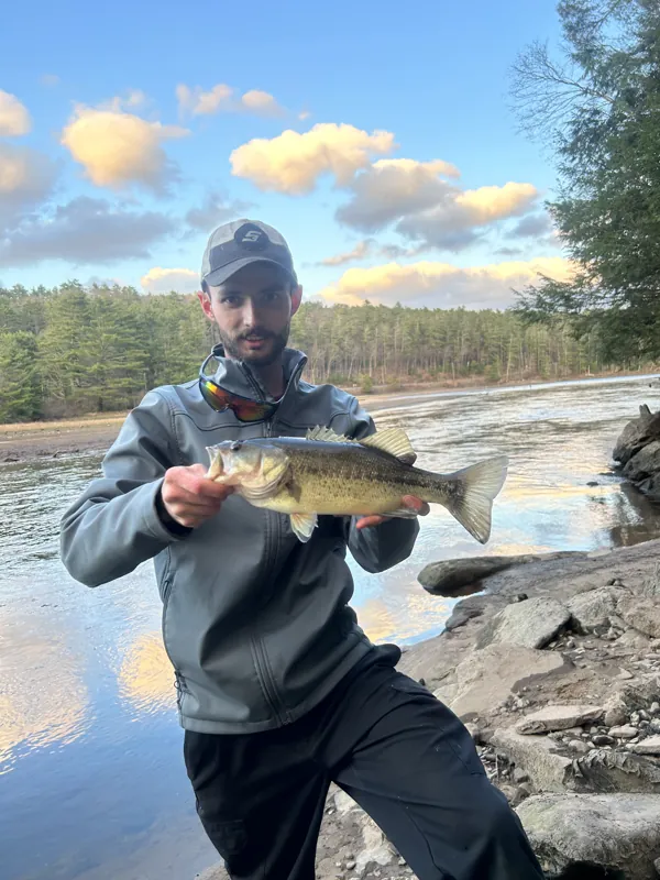An angler kneels on rocks at the water's edge, holding a bass