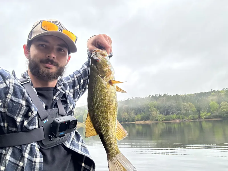 A bearded angler takes a selfie holding up a smallmouth bass over a lake